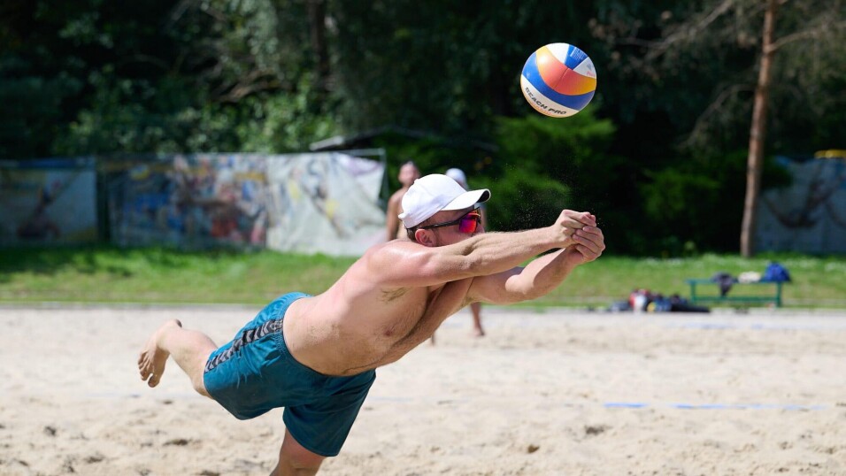 Mężczyzna w&nbsp;białej czapce i&nbsp;okularach rzuca się na&nbsp;piasek, aby odbić piłkę podczas gry w&nbsp;siatkówkę plażową.