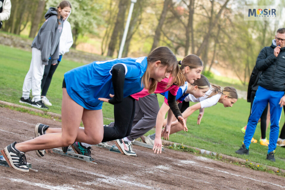 Dziewczęta na&nbsp;blokach startowych gotowe do&nbsp;biegu na&nbsp;bieżni lekkoatletycznej. W tle sędzia z&nbsp;gwizdkiem.