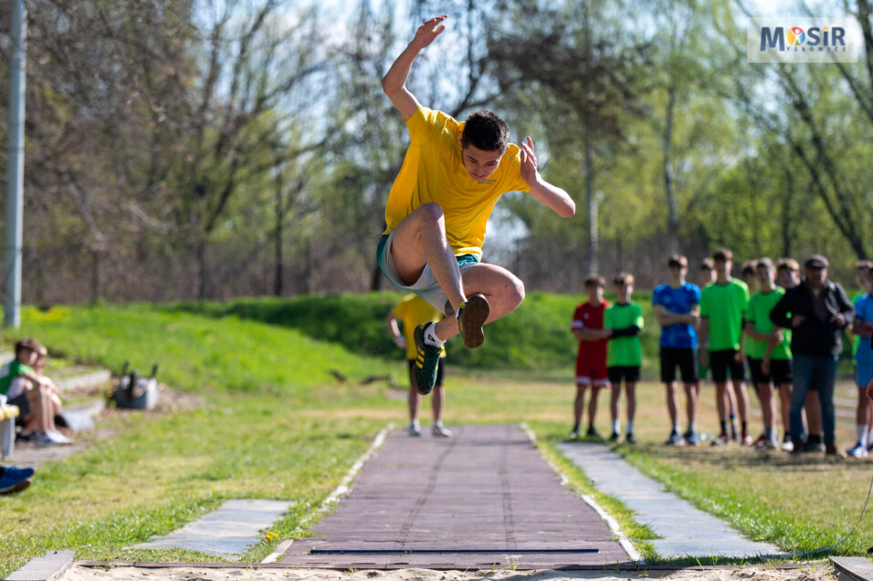 Młody mężczyzna w&nbsp;żółtej koszulce wykonuje skok w&nbsp;dal podczas zawodów. W tle inni uczestnicy i&nbsp;logo MOSiR Mysłowice.