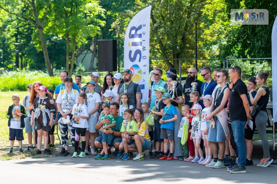 Grupa dzieci i&nbsp;dorosłych z&nbsp;pucharami i&nbsp;medalami pozuje wspólnie w&nbsp;plenerze przy fladze MOSiR Mysłowice.